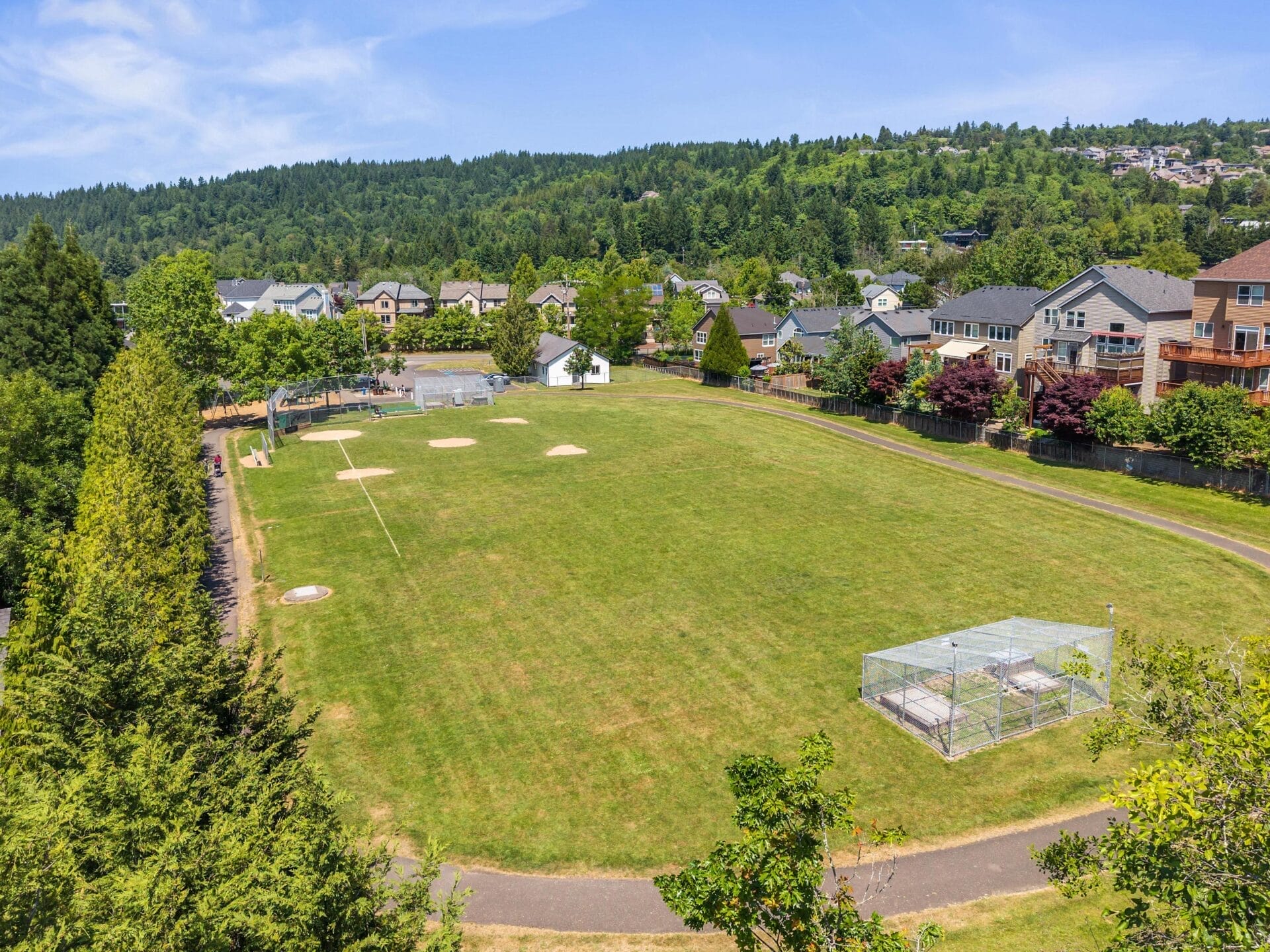Aerial view of a grassy baseball field with bases and a fenced batting cage, surrounded by houses, trees, and hills under a clear blue sky.