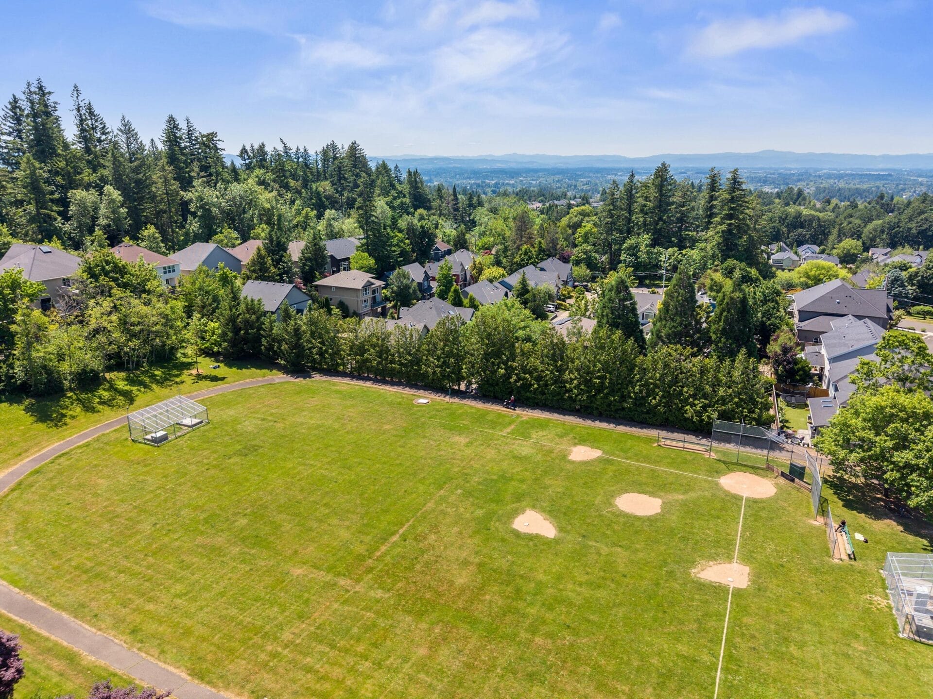 Aerial view of a baseball field with green grass, surrounded by trees and houses, under a blue sky with scattered clouds. The field includes bases, a dirt pitchers mound, and benches.