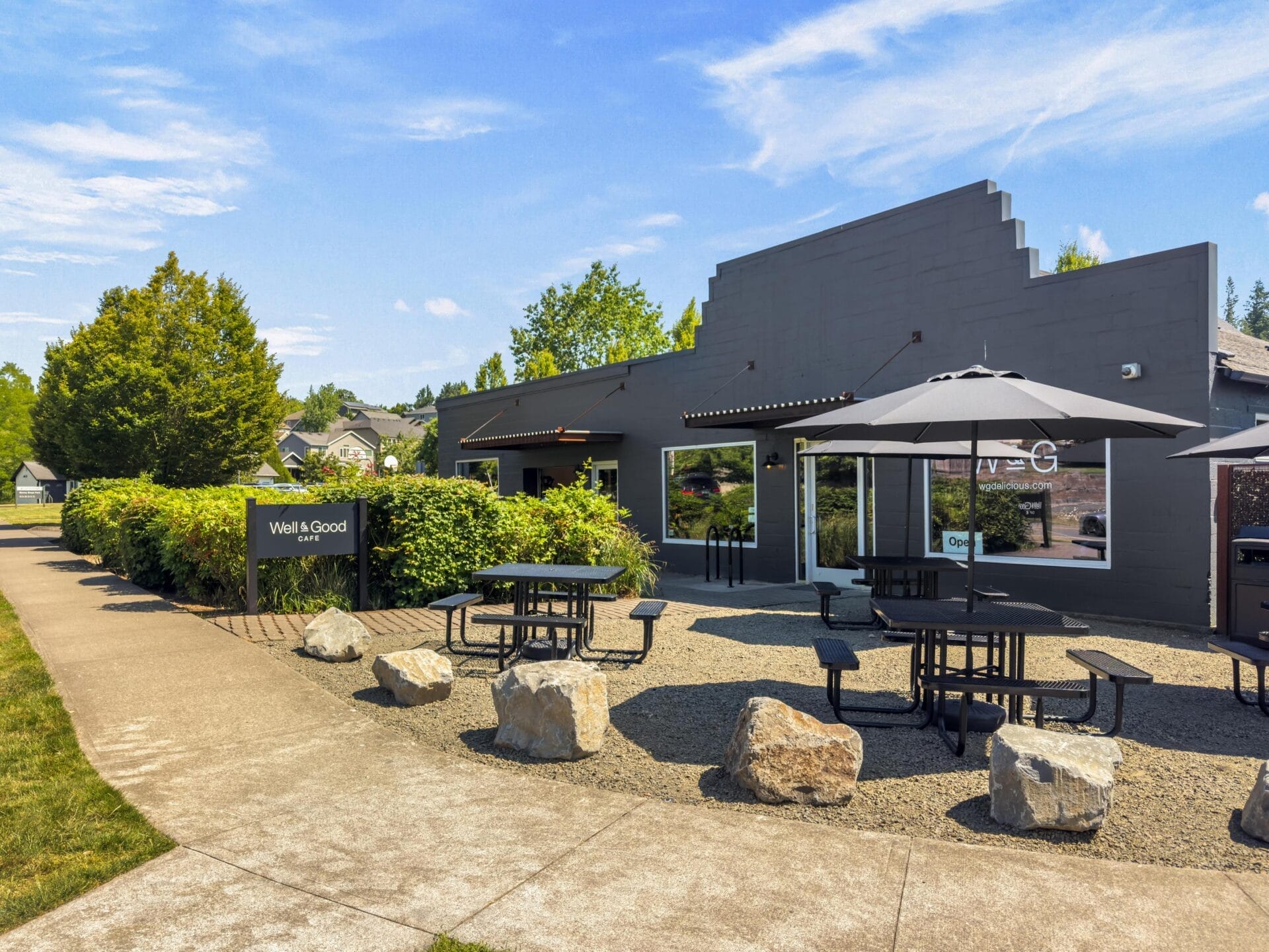 Outdoor seating area with black picnic tables and umbrellas on gravel, surrounded by rocks. Modern dark gray building with large windows and a Well & Good sign. Trees and sidewalk are visible under a blue sky.