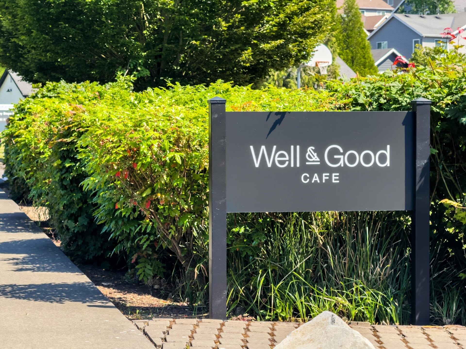 A black sign with white text reading Well & Good Cafe stands in front of green bushes and trees on a sunny day, with part of a sidewalk and houses visible in the background.