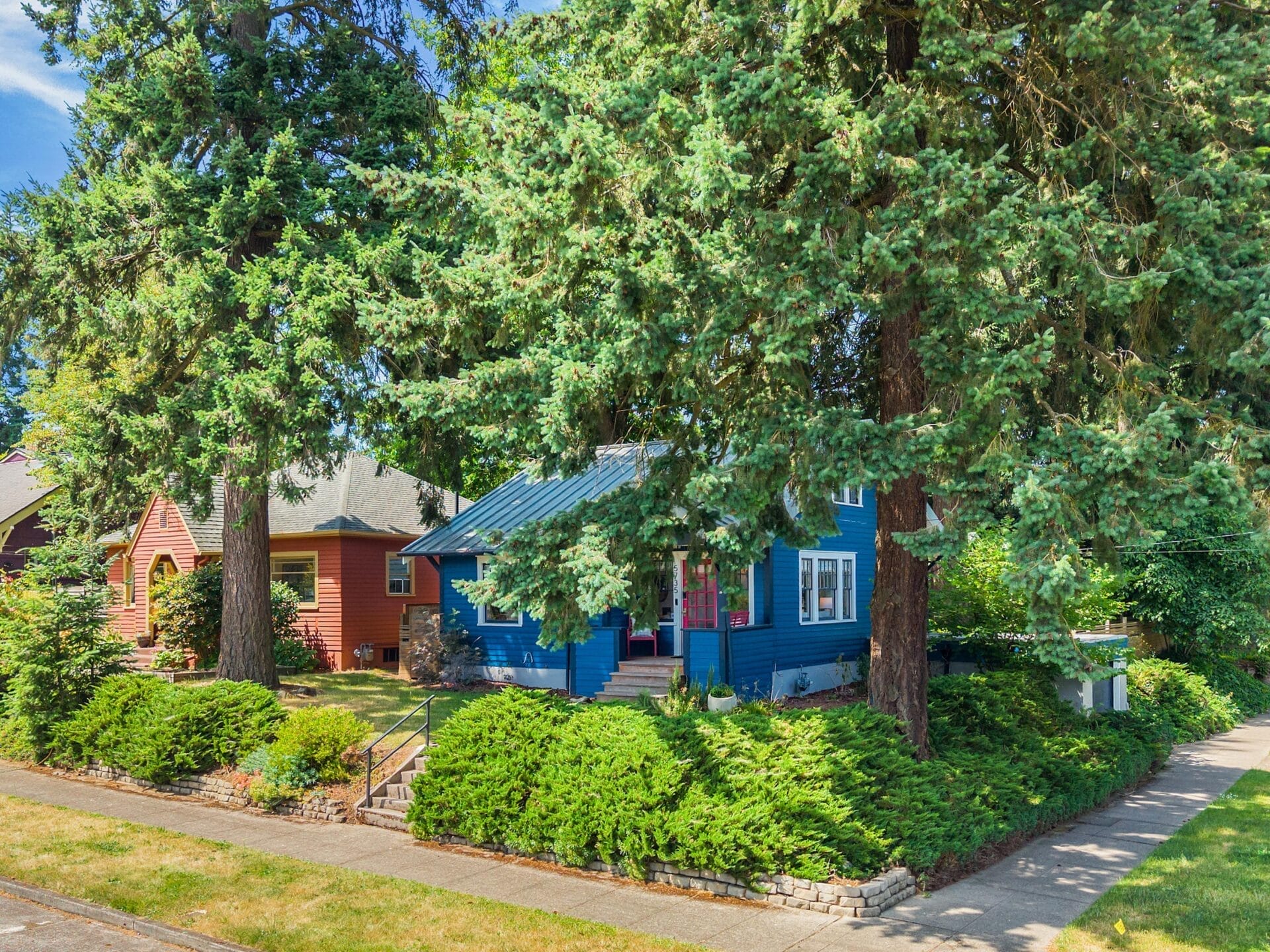 A blue house with a front porch is surrounded by large green trees and bushes on a sunny day in a suburban neighborhood. A red house is partially visible next door. A sidewalk runs along the front of the property.