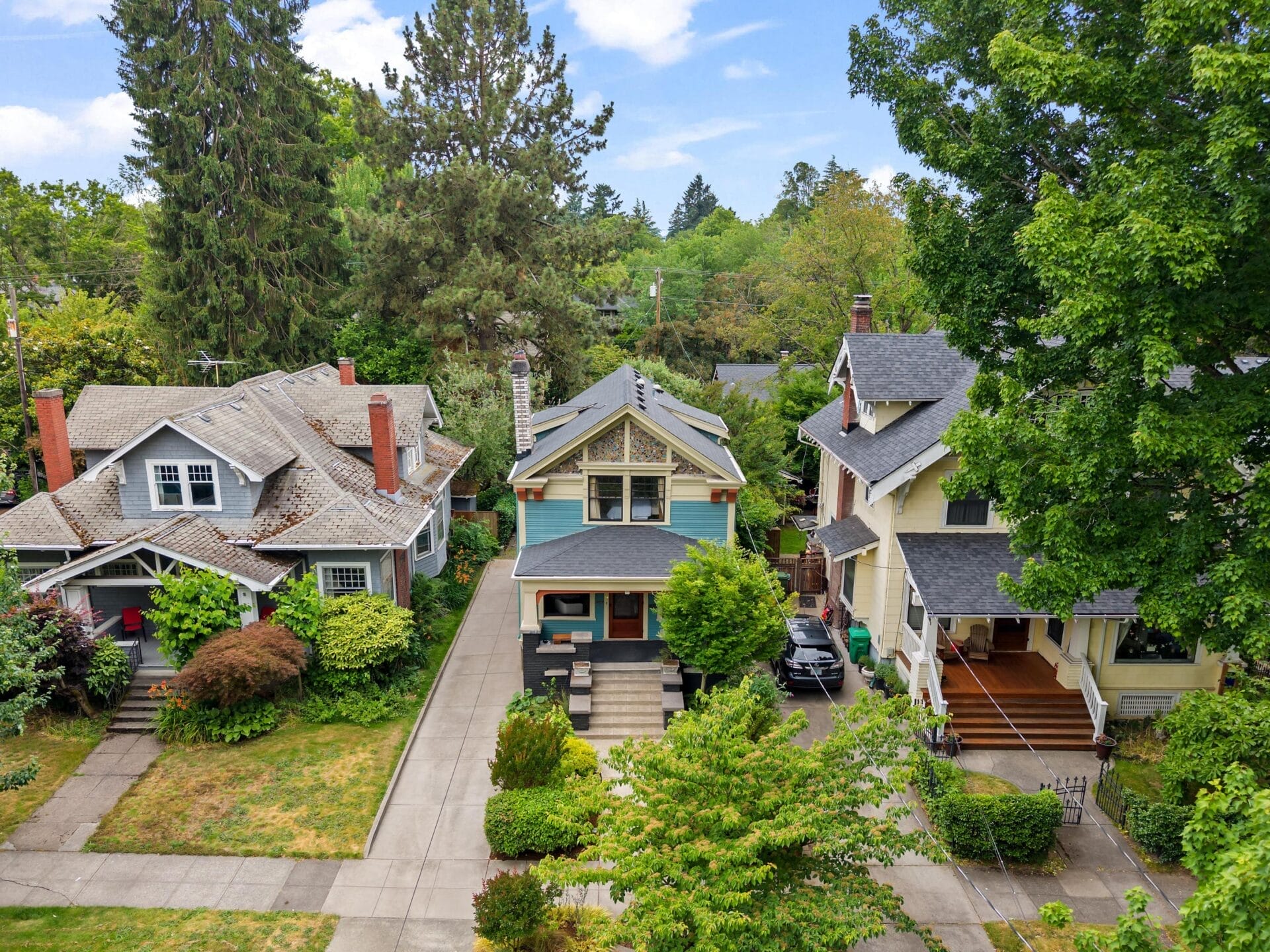 Aerial view of three charming, two-story houses with front porches, surrounded by lush green trees and shrubs on a quiet residential street. The homes have distinct architectural styles and neatly maintained lawns.