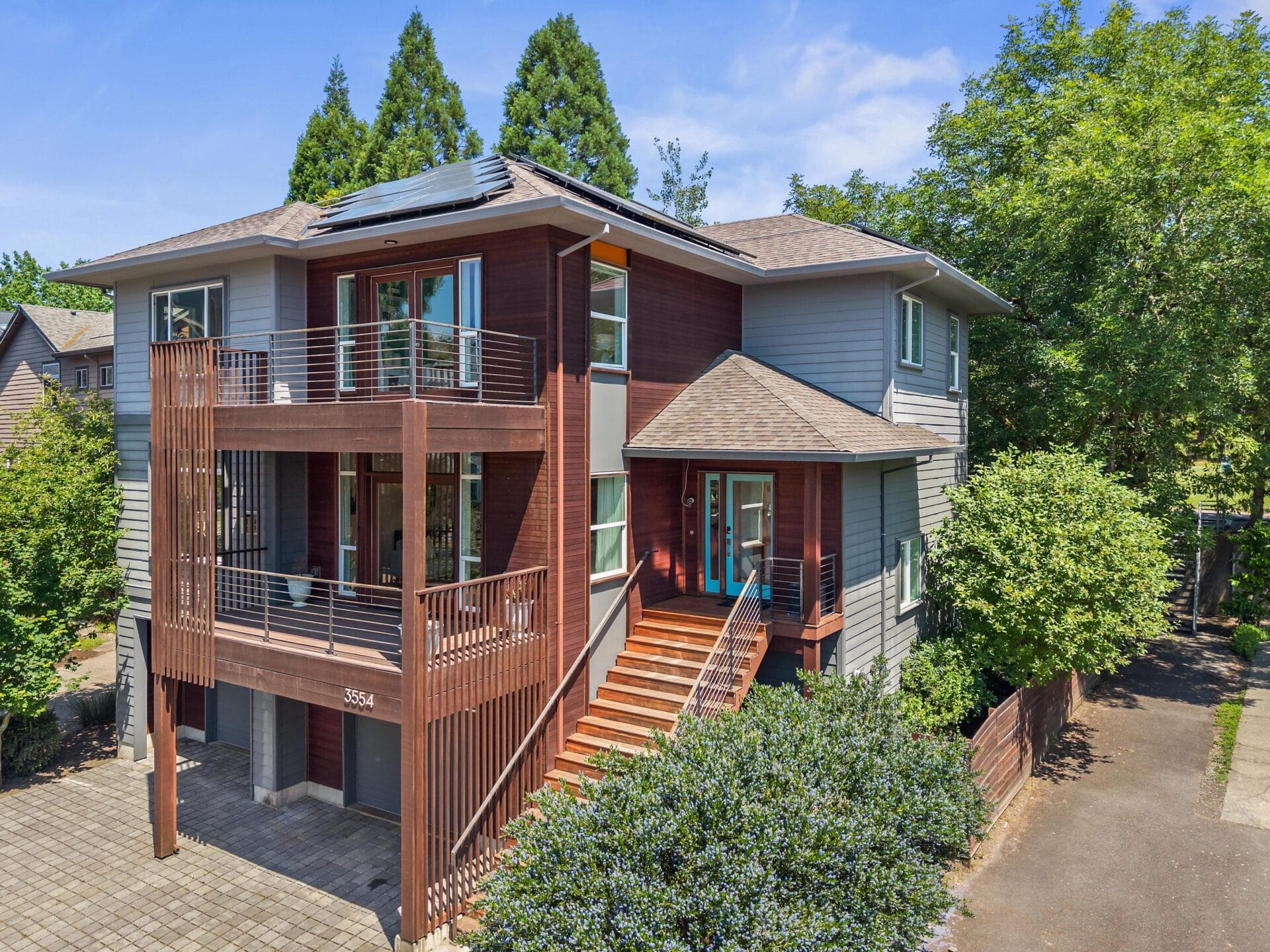 Modern two-story house with brown and gray siding, large windows, solar panels on the roof, and a prominent wooden staircase leading to the front door, surrounded by lush trees and greenery.