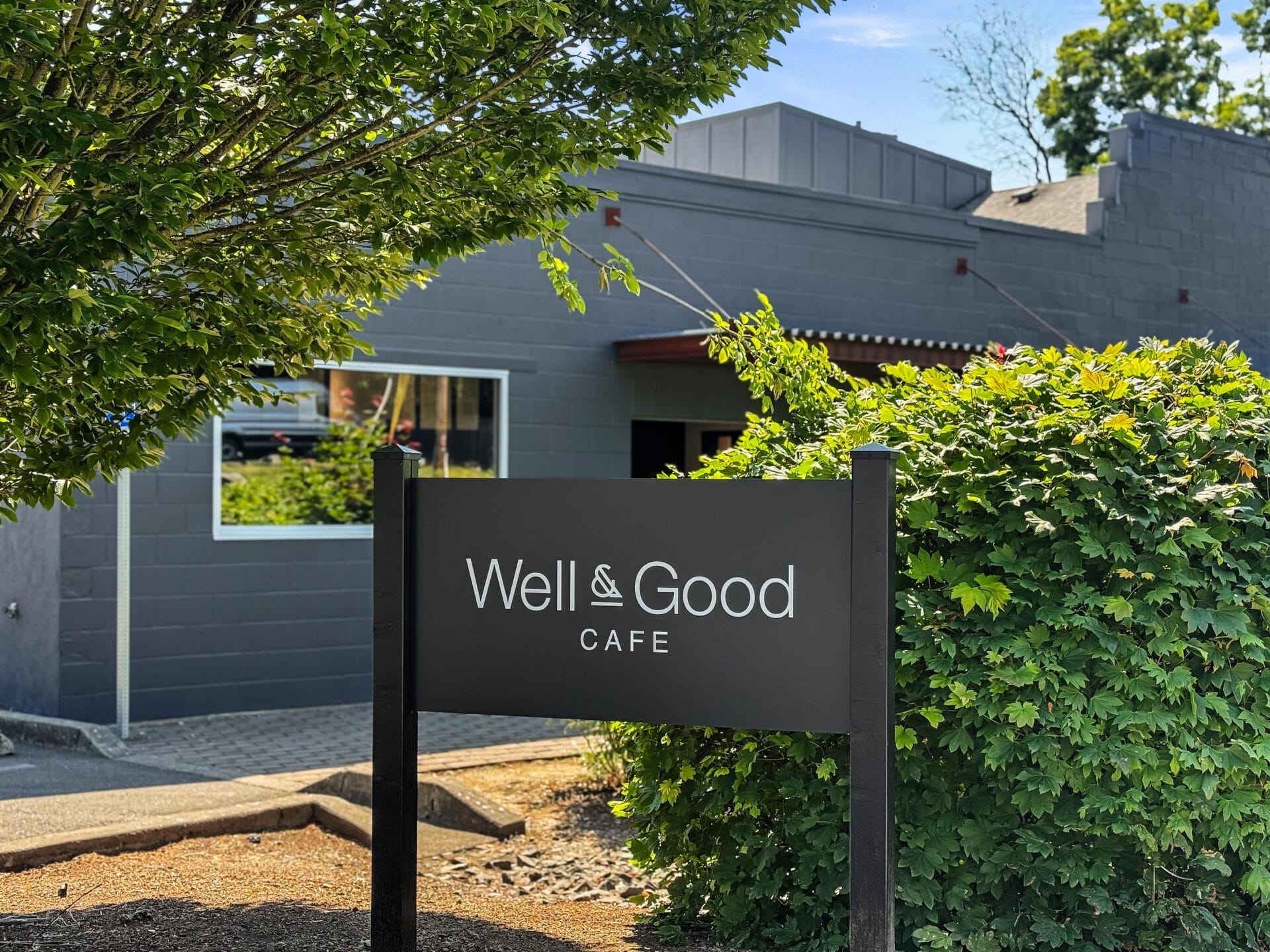 A black sign reading Well & Good Cafe stands in front of a gray building, surrounded by green bushes and trees on a sunny day.