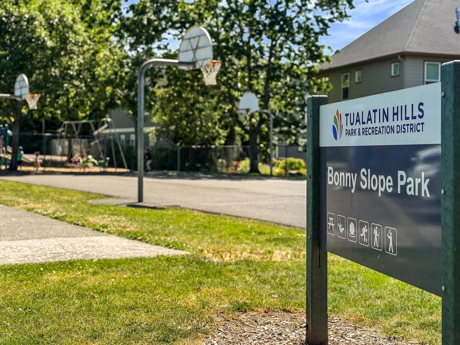 A sign for Bonny Slope Park in the Tualatin Hills Park & Recreation District stands in front of basketball hoops, a playground, and trees on a sunny day.