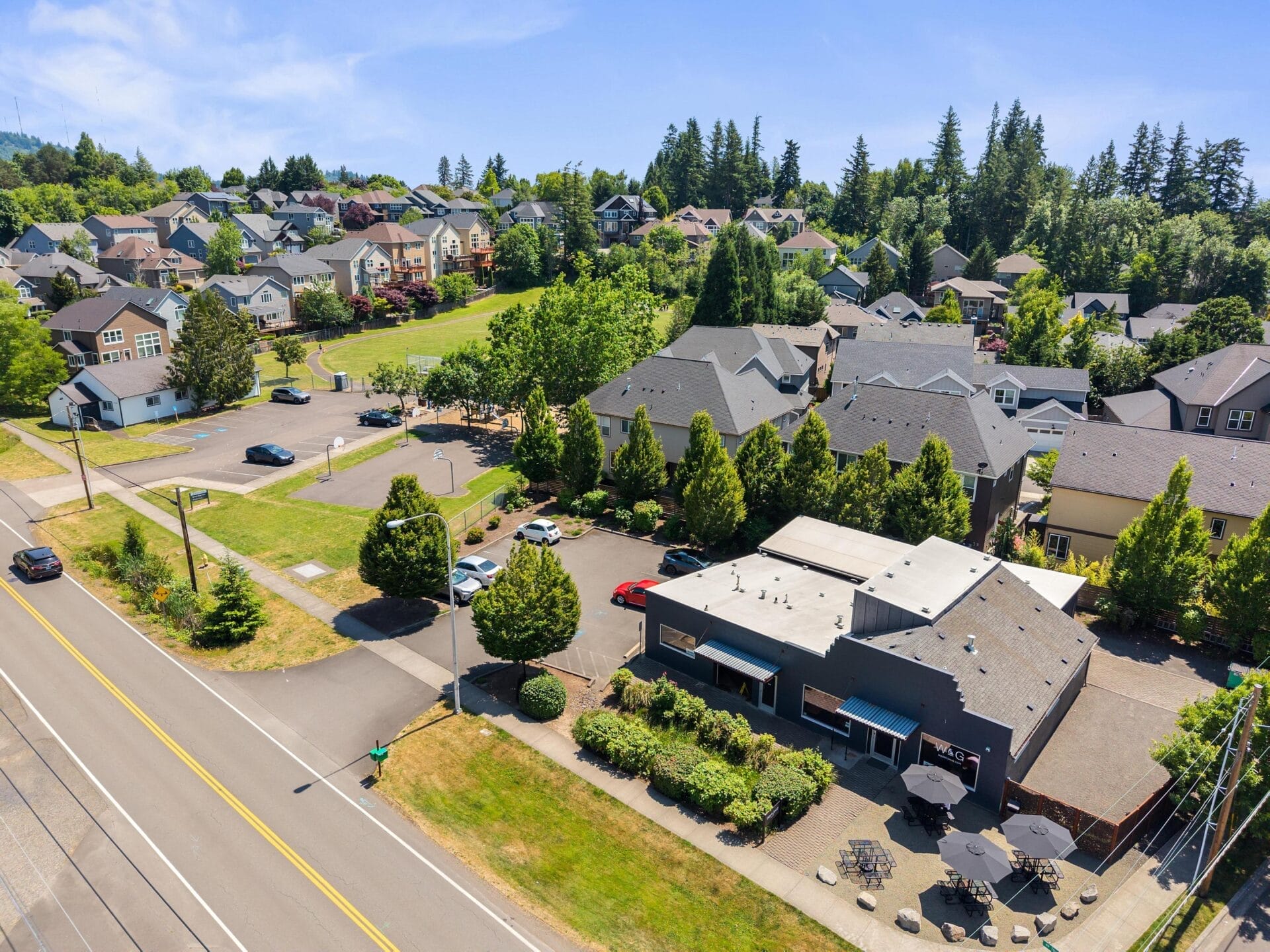 Aerial view of a suburban neighborhood with houses surrounded by trees, a small commercial building with outdoor seating, parked cars, and roads, all under a clear blue sky.