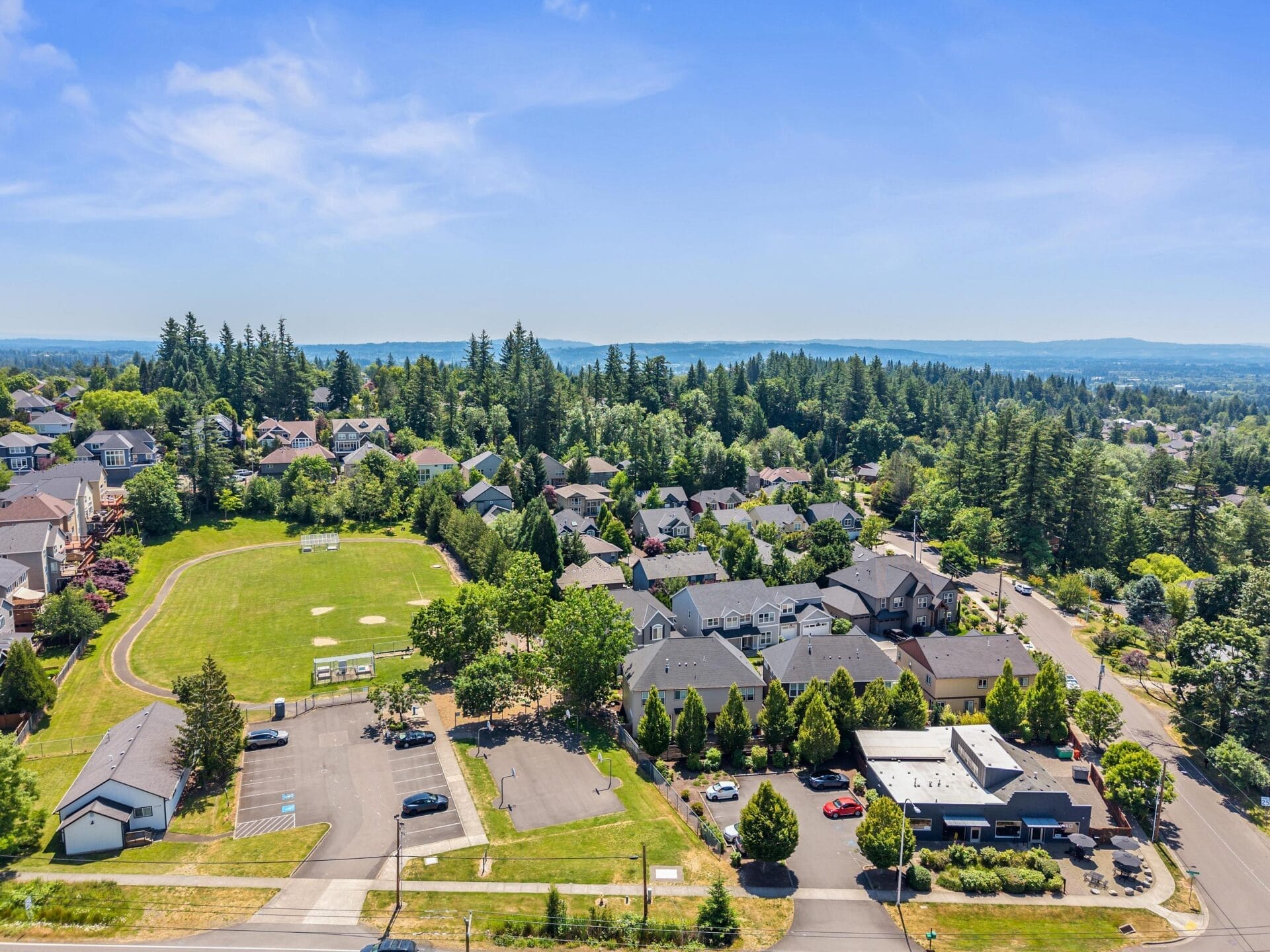 Aerial view of a suburban neighborhood with houses surrounded by trees, a green sports field with soccer goals, parking areas, and clear blue sky in the background.