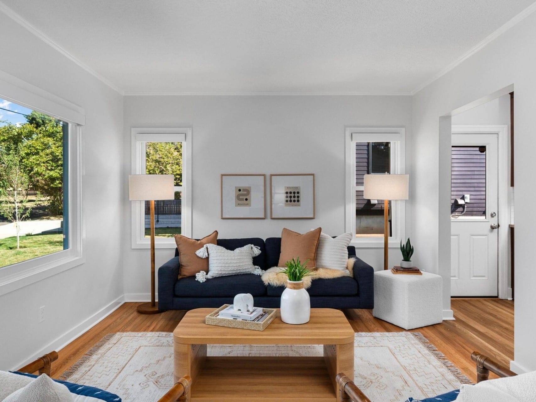 A bright living room with large windows, a dark blue sofa with pillows, two standing lamps, a wooden coffee table with decor, and light walls and wooden flooring. Natural light fills the cozy, modern space.