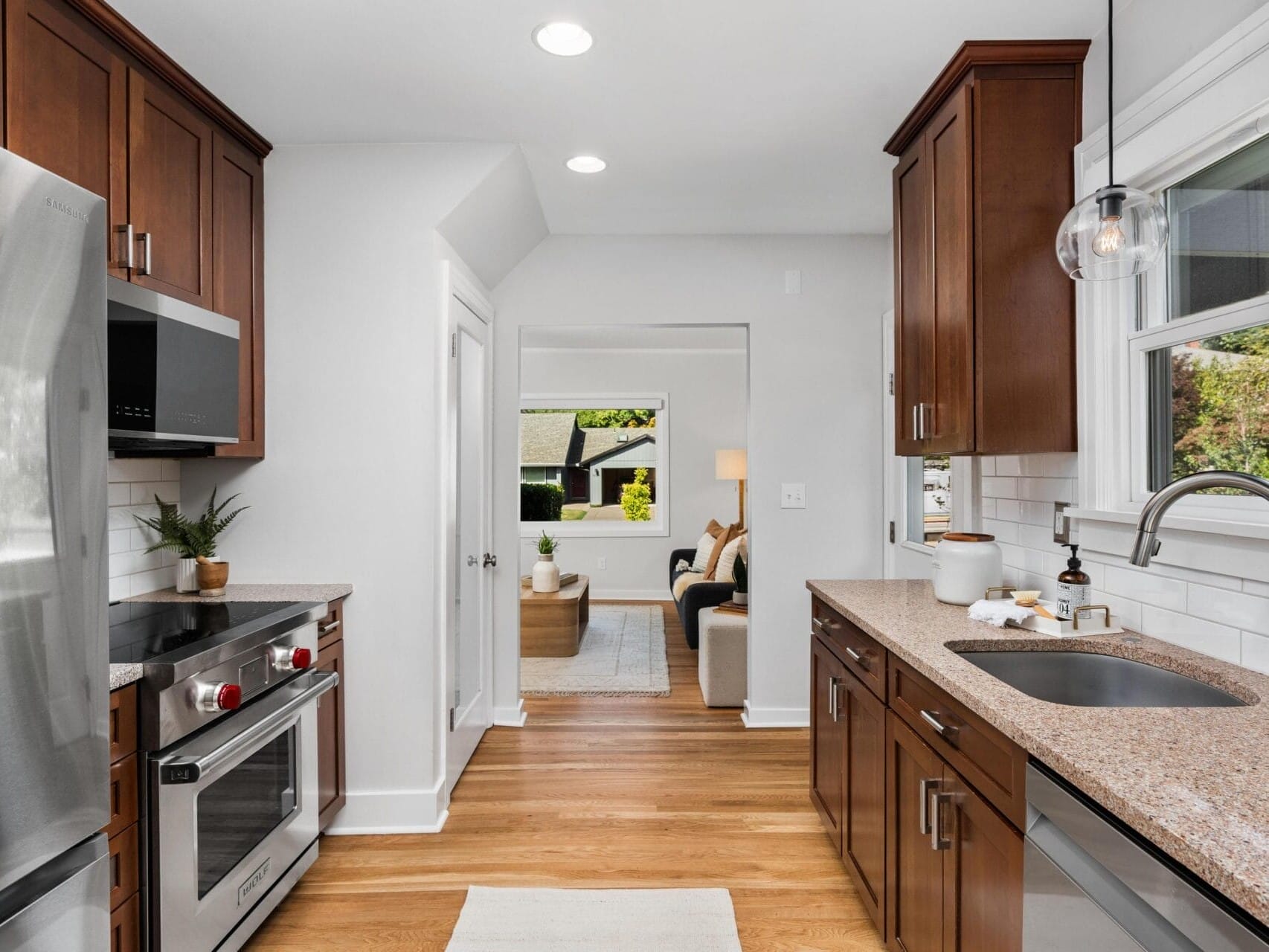 Modern kitchen with stainless steel appliances, brown cabinets, granite countertops, and a large window above the sink. The space opens to a bright living area with a sofa and greenery visible outside.