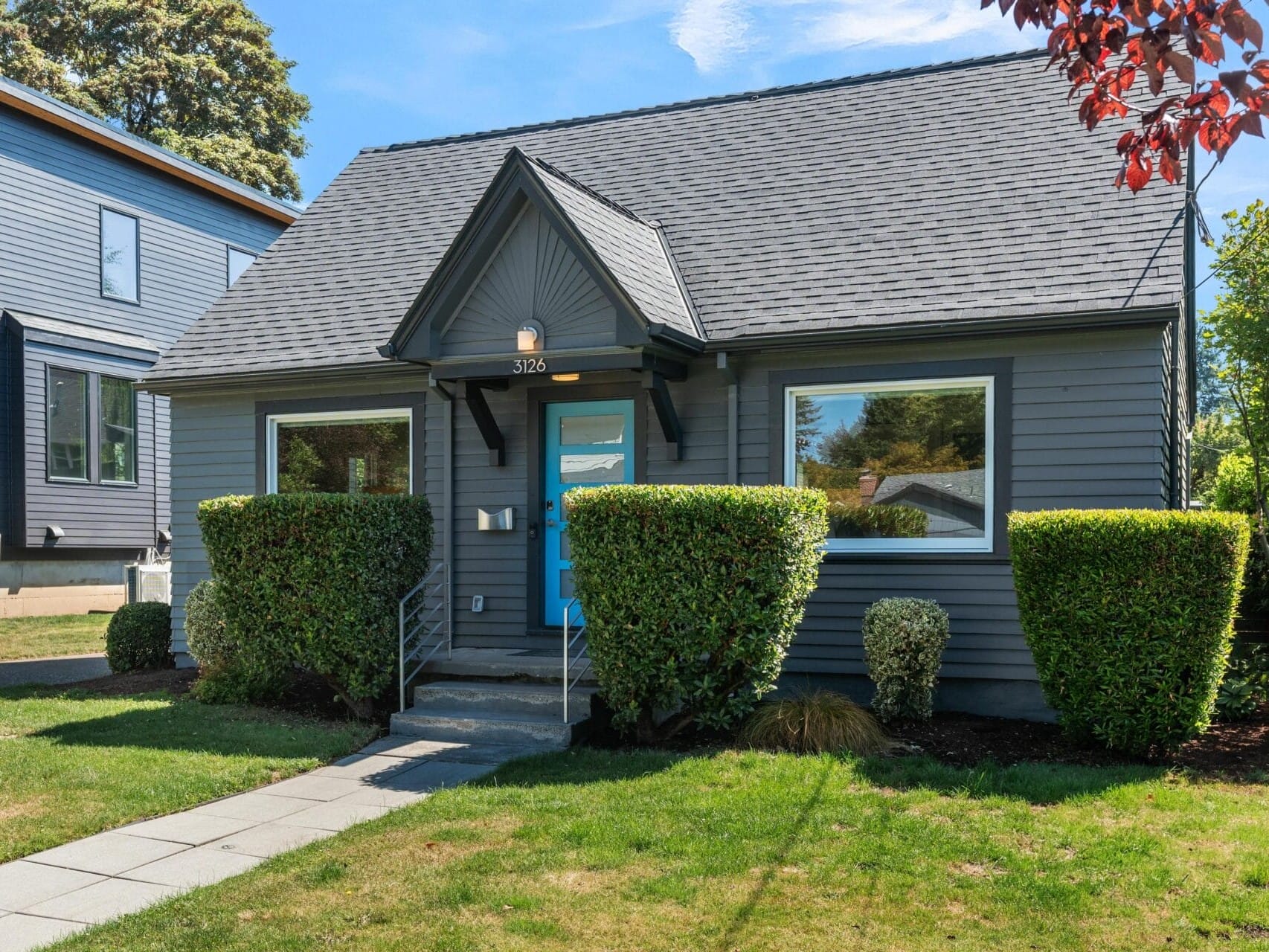A small gray house with a steep, dark roof and bright blue front door, surrounded by neatly trimmed bushes and a green lawn, with a concrete walkway leading to the entrance.