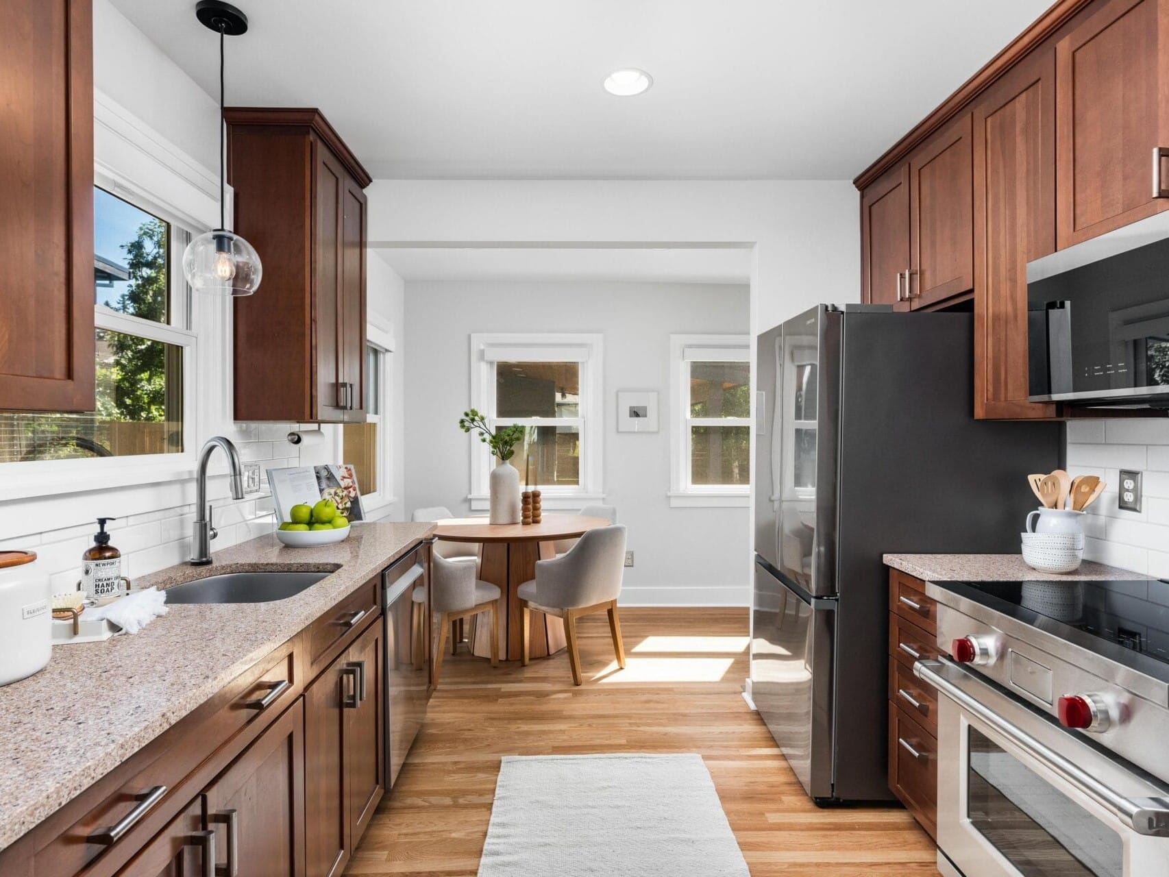 Modern kitchen with wood cabinets, stainless steel appliances, granite countertops, and a small dining area with a round table and two chairs by windows; bright natural light fills the space.