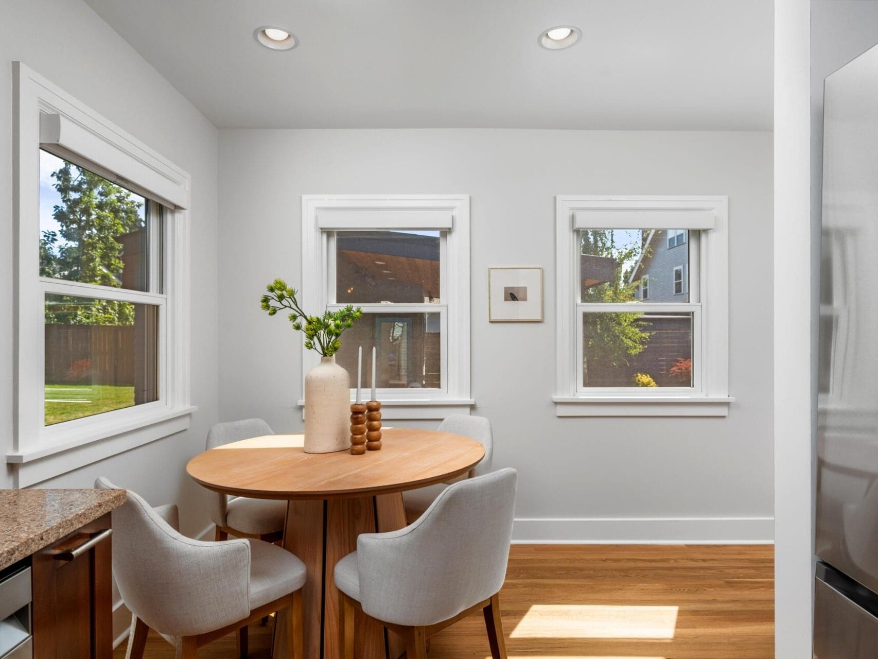 A small dining area with a round wooden table, four upholstered chairs, and two tall vases with greenery, set beside three windows letting in natural light. The room has light walls and a wooden floor.