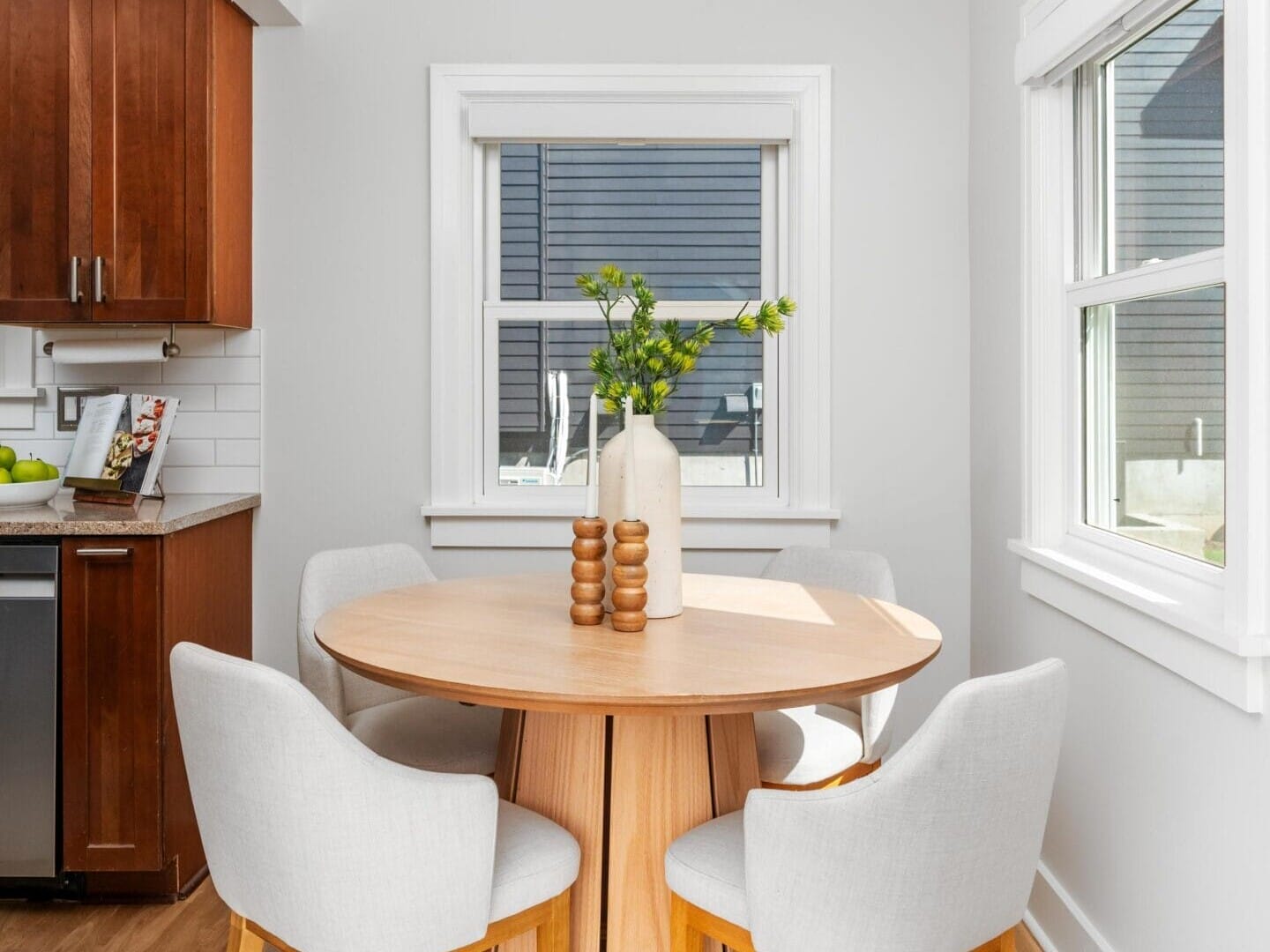 A small round wooden dining table with four light gray upholstered chairs is set in a bright kitchen nook. Two white vases with green branches sit on the table, and sunlight streams in through nearby windows.