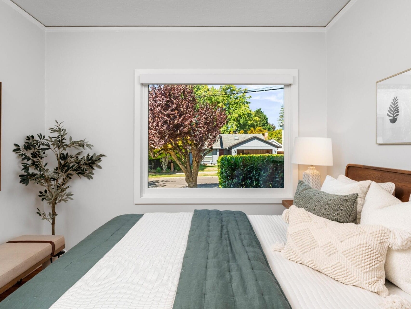 A neatly made bed with green and white bedding is in a bright bedroom. There is a large window showing a tree and suburban houses outside. The room features a plant, artwork, a bench, and a bedside lamp.