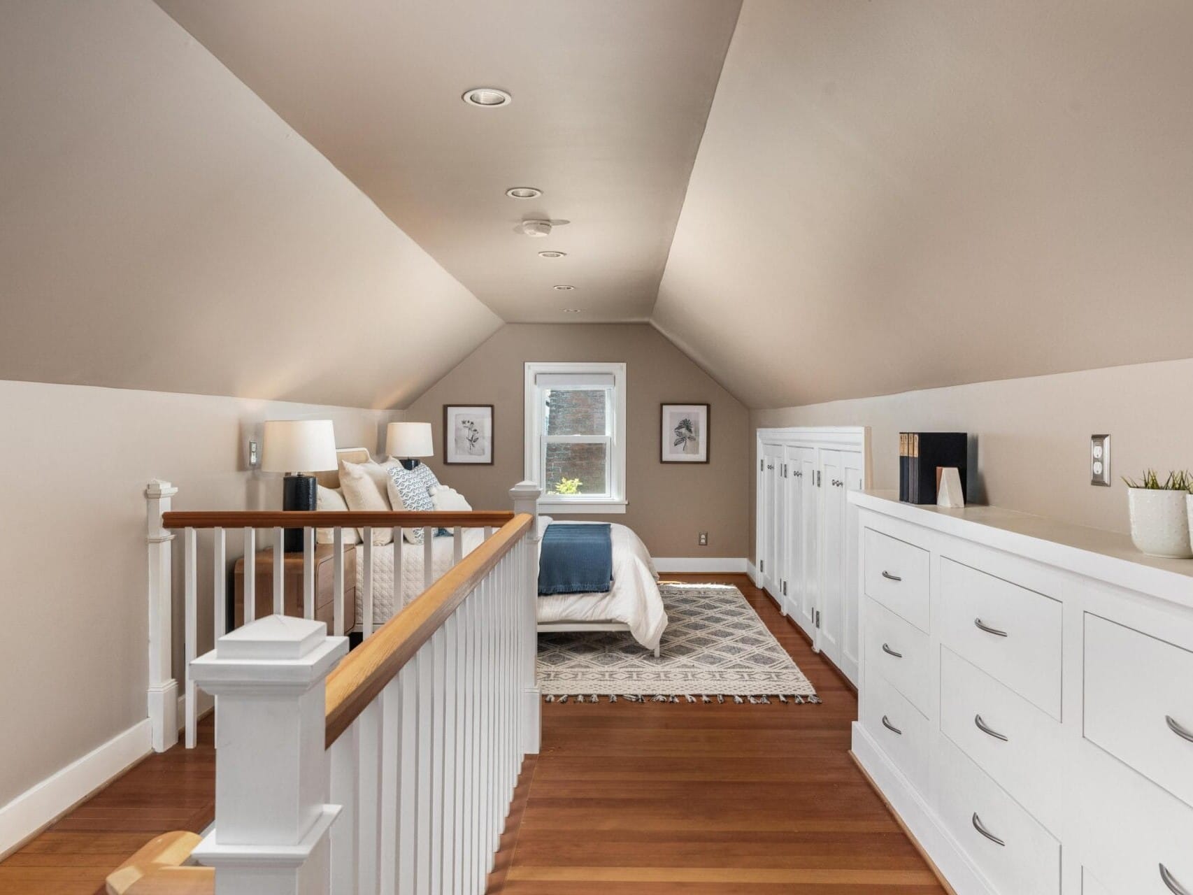 A cozy attic bedroom with sloped ceilings features a bed with white bedding, nightstands with lamps, framed wall art, a small window, wooden floors, and built-in white drawers along the right wall.
