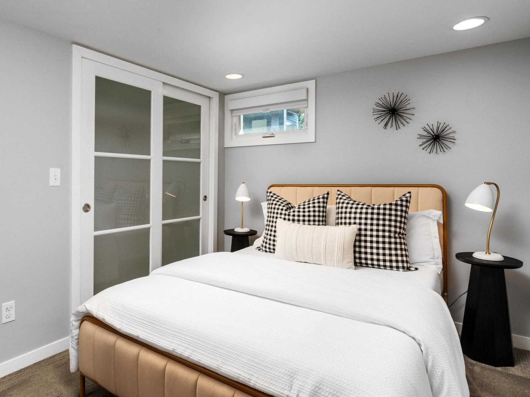 A modern bedroom with a neatly made bed, neutral bedding, plaid pillows, two small black side tables with lamps, wall art above the headboard, gray walls, and a sliding door closet.