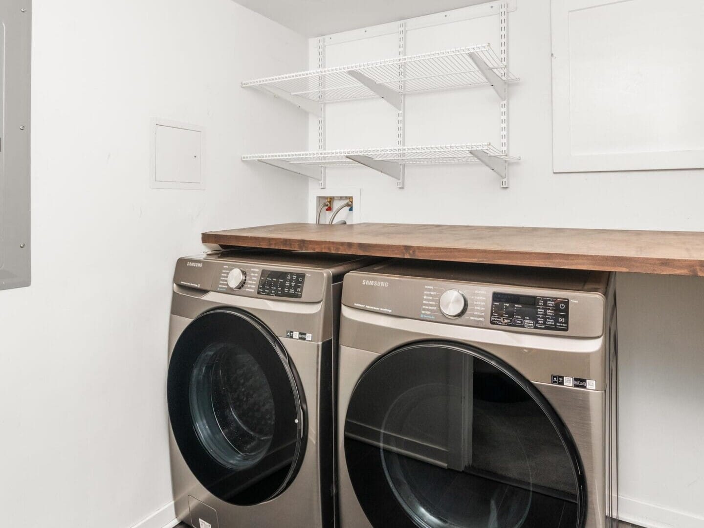 A modern laundry room with a front-loading washer and dryer set under a wooden countertop, white walls, gray wood flooring, and white metal shelves mounted above.