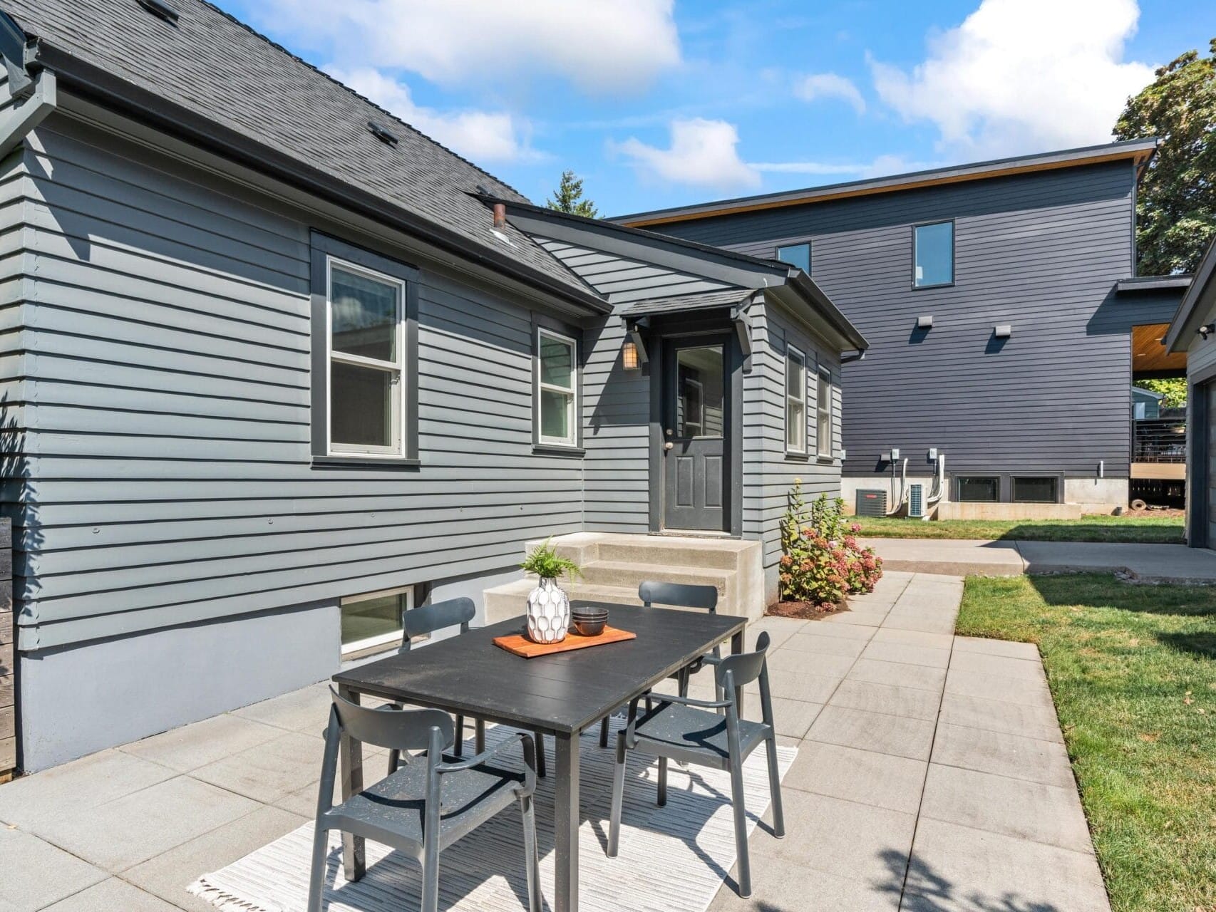 A modern patio with a black outdoor dining table and four chairs on a rug, set beside a gray house with siding. There is a small lawn, nearby shrubs, and a larger dark building in the background under a blue sky.