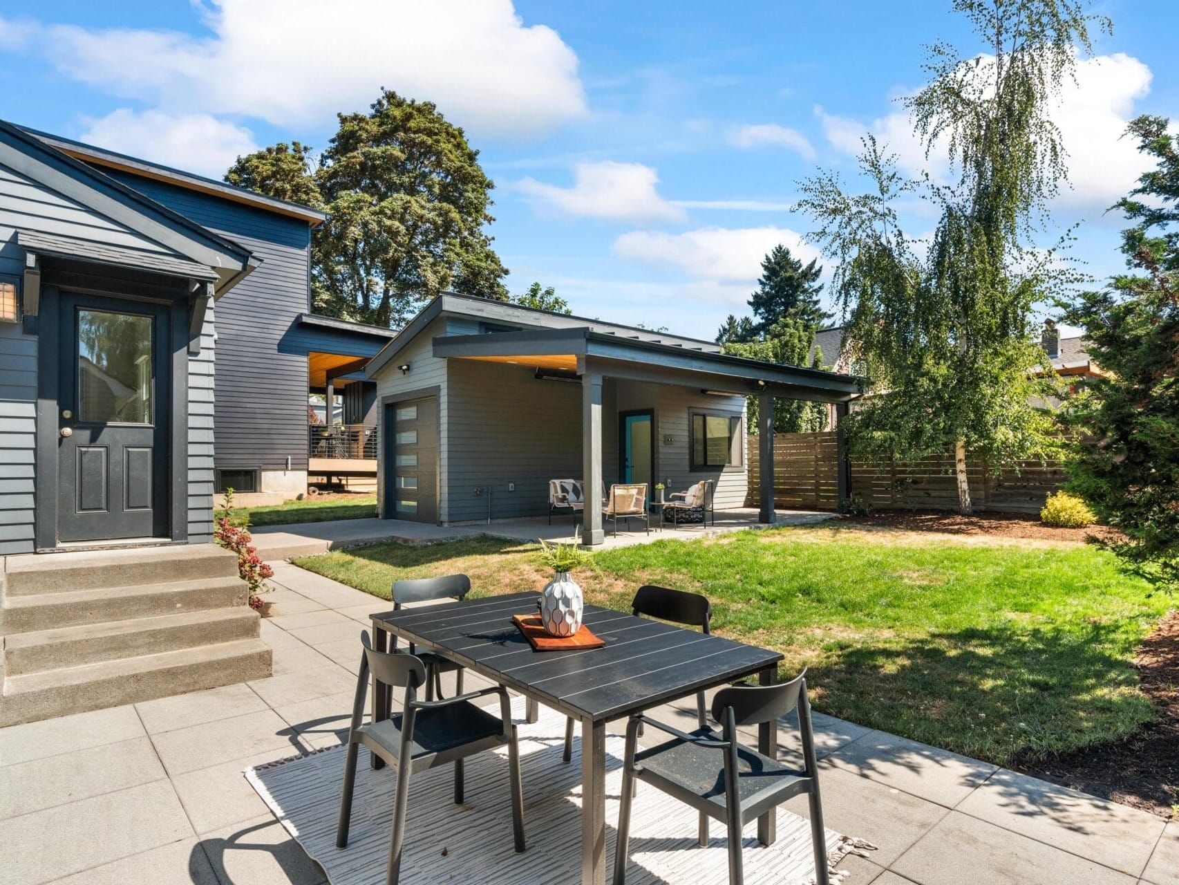 Modern backyard with a patio featuring a black outdoor dining set on a rug, steps leading to a gray house, green lawn, trees, and a detached covered seating area under a blue sky with scattered clouds.