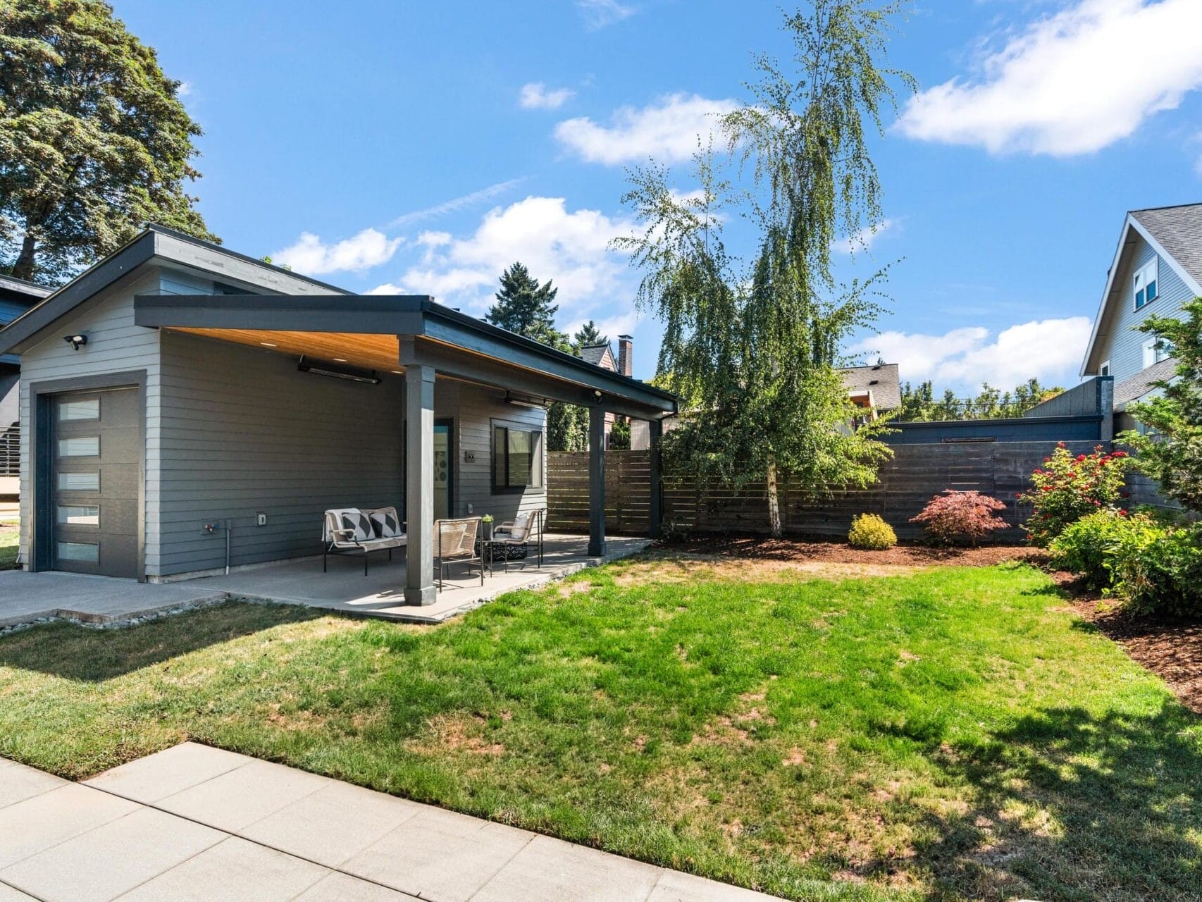 A modern house with a covered patio, outdoor seating, and a well-maintained backyard lawn. There are trees, shrubs, and a wooden fence surrounding the yard under a partly cloudy sky.