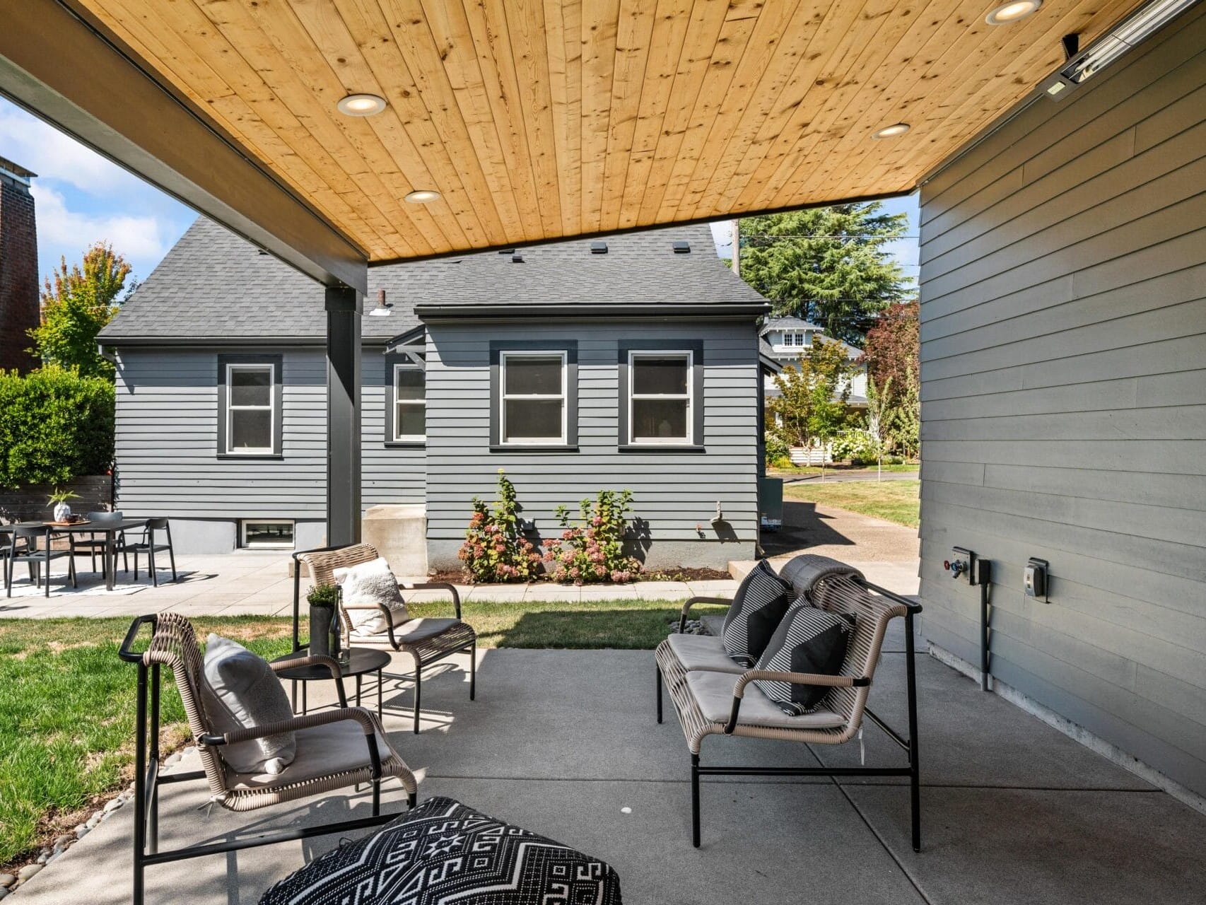 A covered patio with cushioned chairs and a small table looks out onto a backyard and a gray house with white trim on a sunny day. Patio lights are installed in the wooden ceiling above.