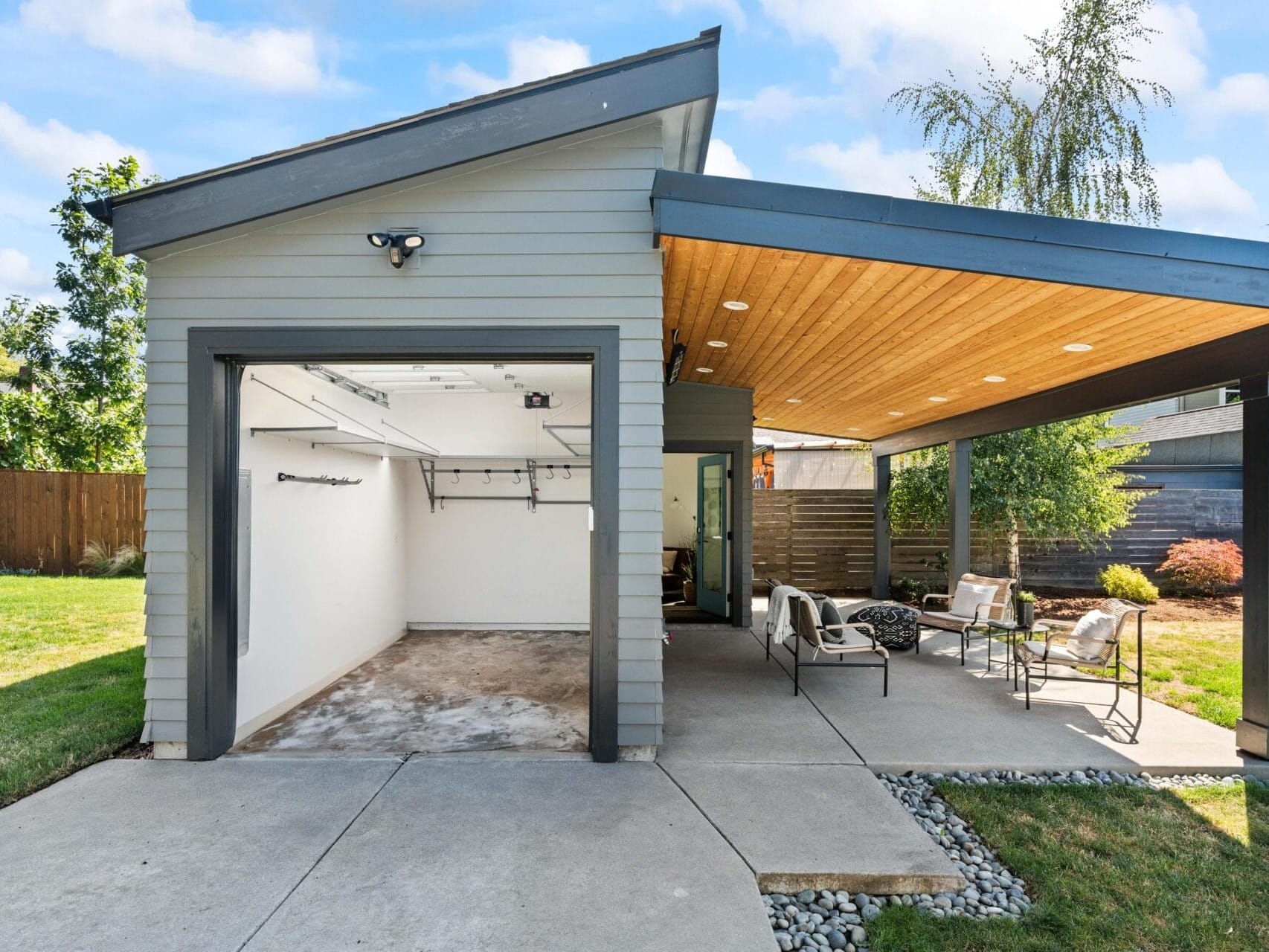 A modern outdoor patio with a wooden ceiling is attached to a small gray garage. The covered area has chairs, a table, and seating, and is surrounded by green grass and a wooden fence on a sunny day.