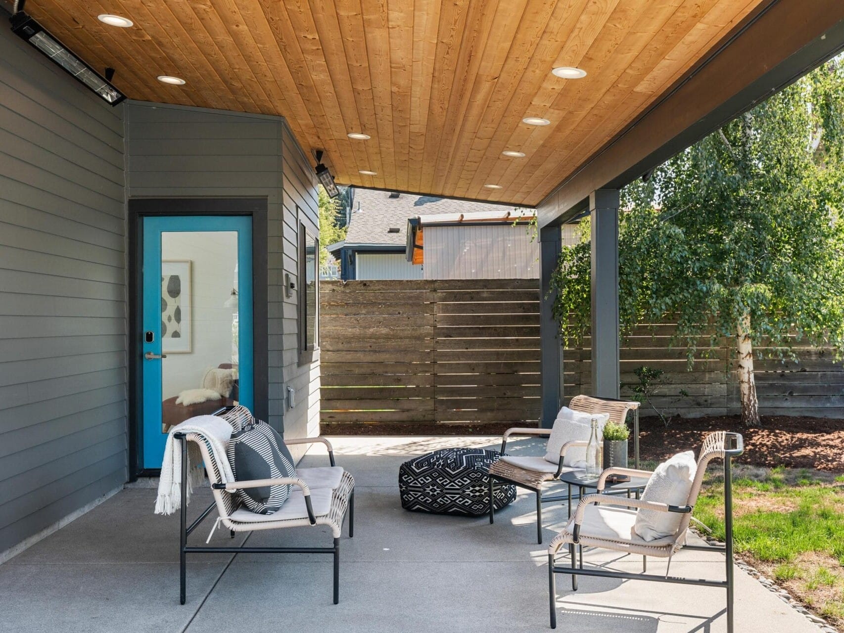 A modern covered patio with a wooden ceiling, gray siding, patio chairs with cushions and blankets, a round patterned ottoman, and a blue-trimmed door leading inside. A wooden fence and trees are in the background.
