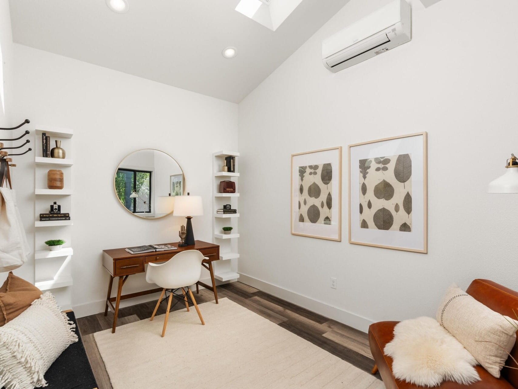 A modern, minimalist home office with a wooden desk, round mirror, white chair, wall art, shelves with decor, a tan rug, a brown leather chair with pillows, and a bench with a pillow and bag under white walls and ceiling lights.