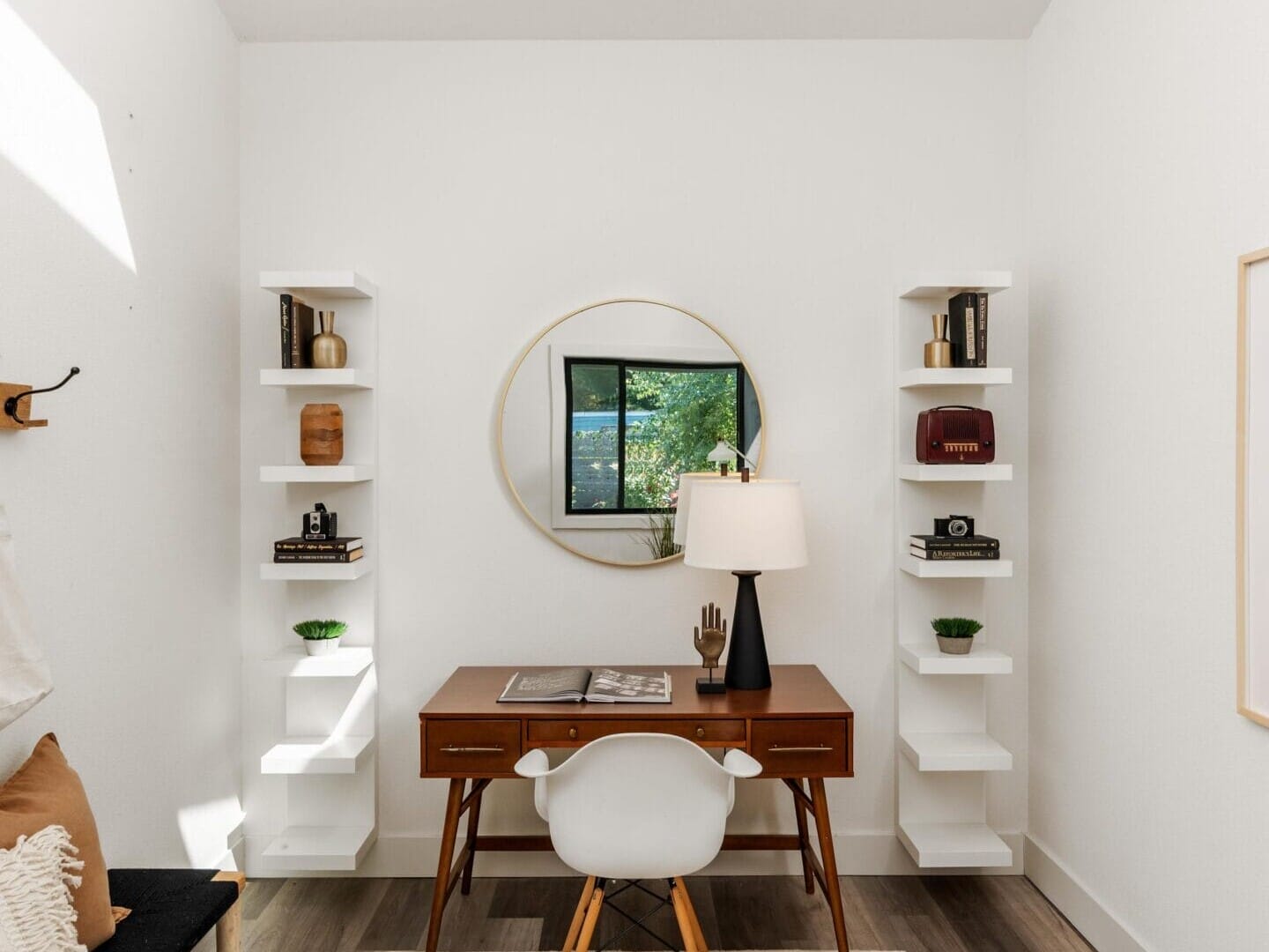 A modern, minimalist home office with a wooden desk, white chair, lamp, and round mirror. Floating shelves hold decor items. A window shows greenery outside. Neutral tones and a rug complete the space.