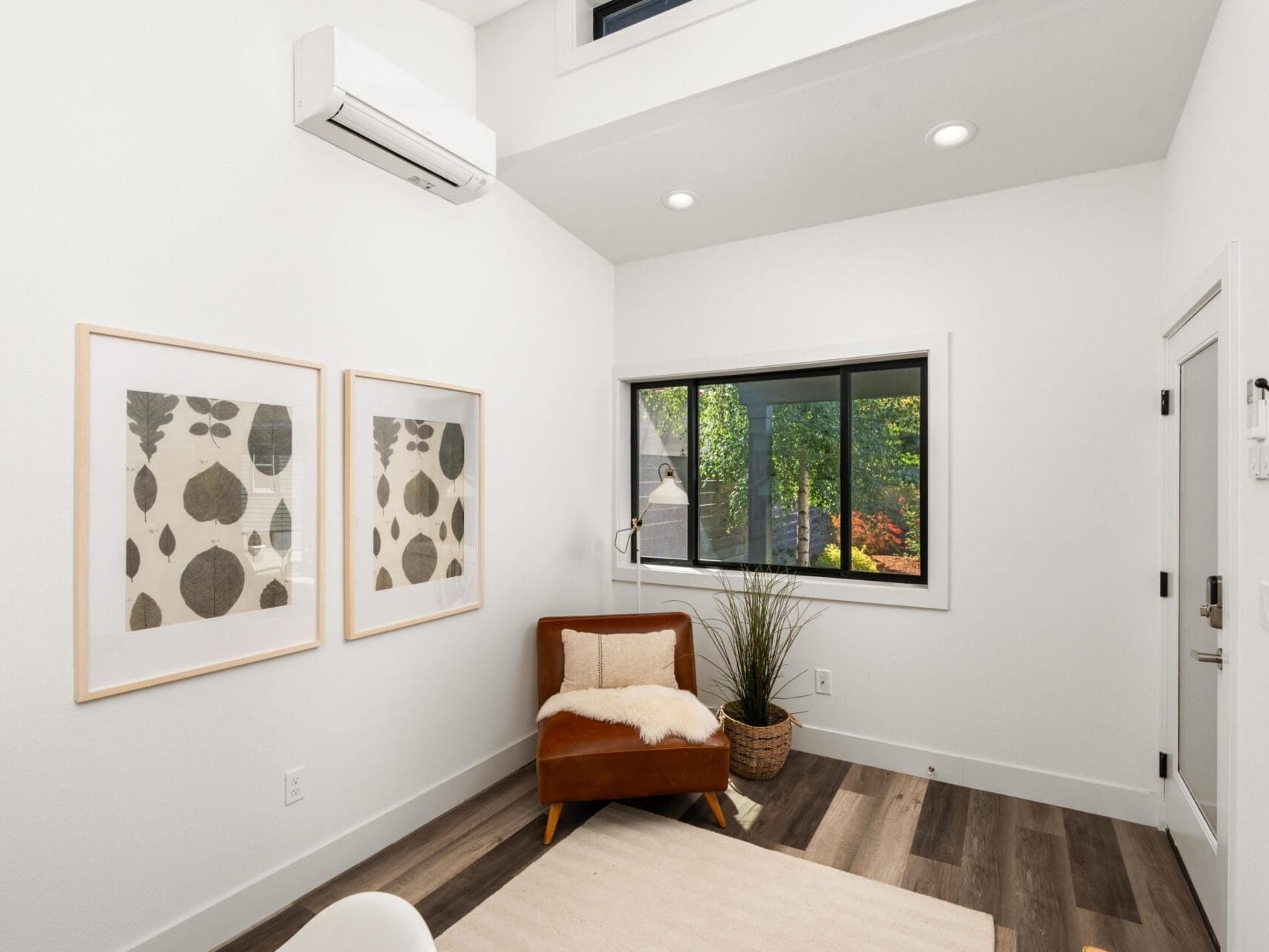 A small, modern room with white walls, wood flooring, a brown accent chair with a pillow and throw, two framed abstract prints, a window with a view of greenery, and a potted plant in the corner.