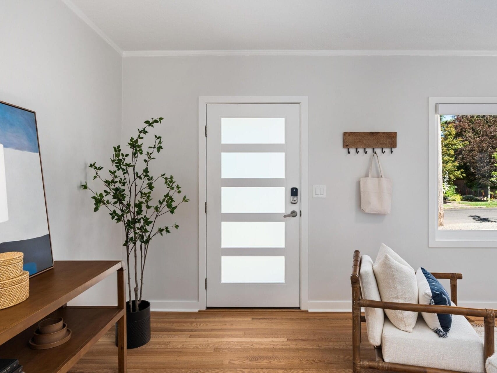 A modern entryway with a white door featuring frosted glass panels, a wooden bench with cushions, a small plant, a wall hook with bags, and a console table with decor, all on a hardwood floor.
